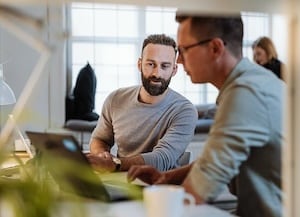 two people sitting beside each other at a desk looking at laptop