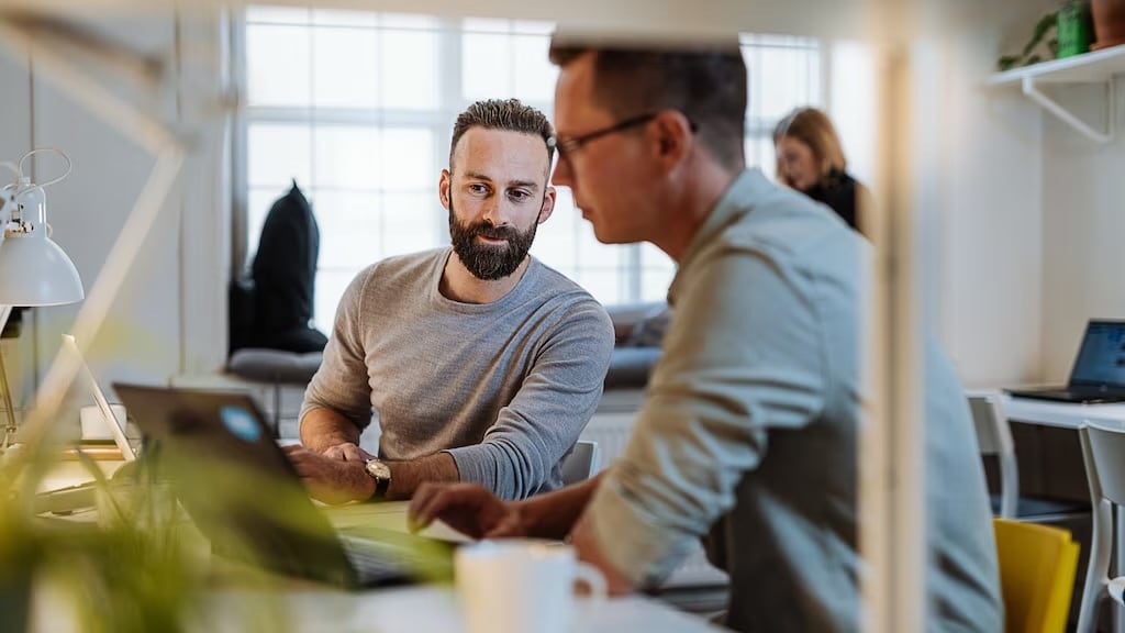Two individuals sitting at desk next to each other looking at laptop