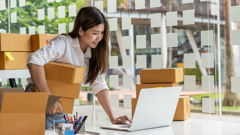 Woman standing at desk looking at laptop and holding boxes
