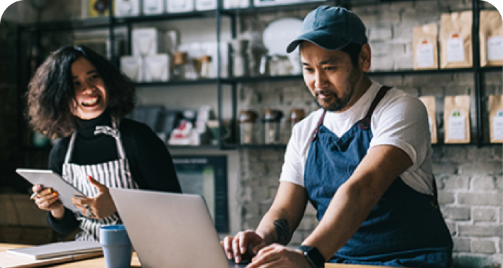 Two coffee shop owners sittng behind counter and managing business on laptop and tablet