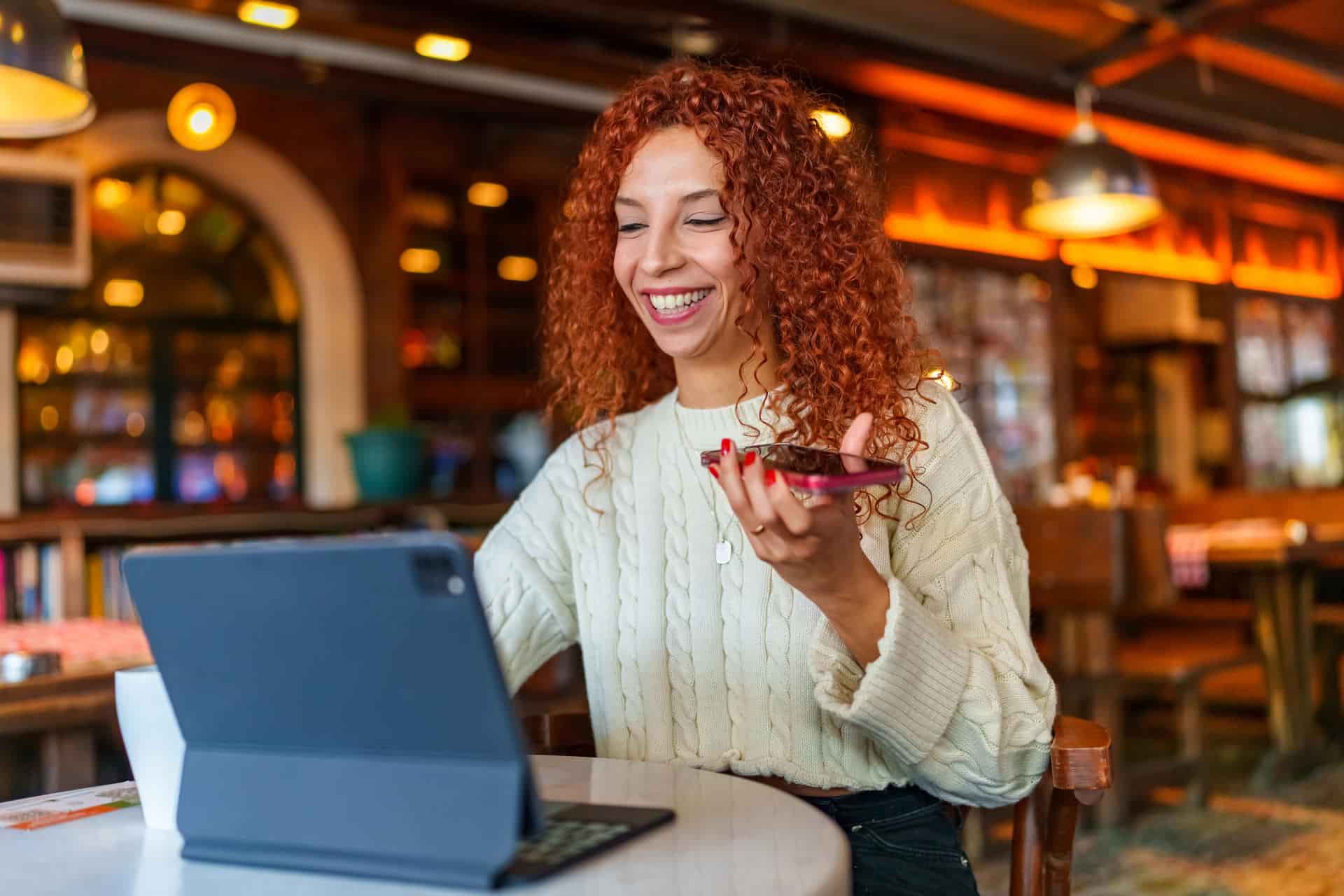 Frau mit roten und gelockten haaren redet am Telefon während sie vor einem Laptop im Cafe sitzt.