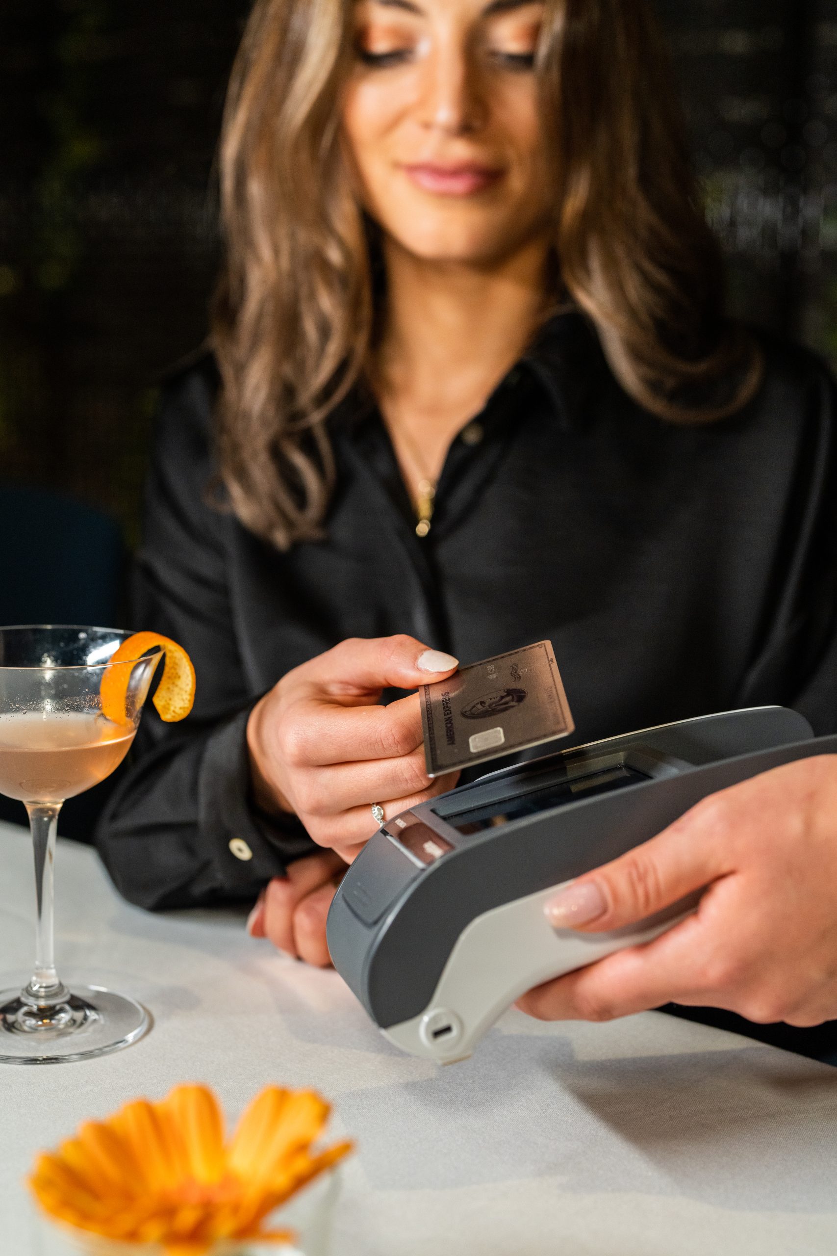 Eine junge Frau hält am Tisch eines Restaurants eine AMEX Gold Card an ein Lesegerät. Neben ihr ein Cocktailglas.