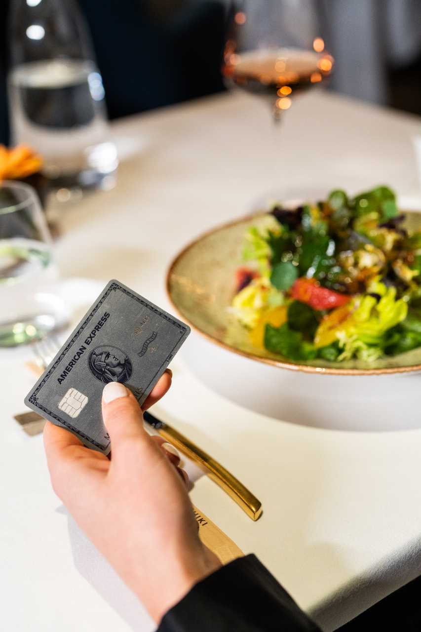 Person hält eine silberne AMEX Kreditkarte bereit zum Zahlen. Im Hintergrund ist ein essen mit Weinglas am Tisch serviert.