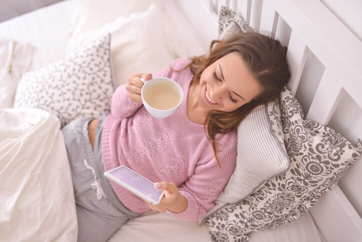 Frau liegt mit einem Kaffee in der rechten und einem Tablet in der Linken Hand im Bett