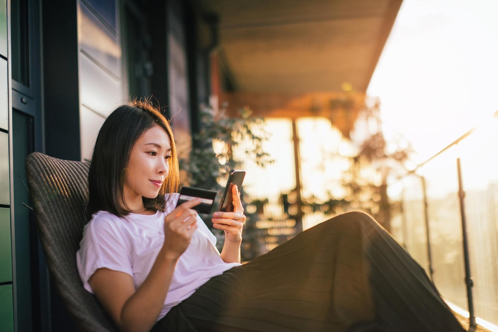 Frau sitzt in einem Sessel auf einer Veranda, in der linken Hand ihr Handy und in der rechten Hand eine Kreditkarte haltend.