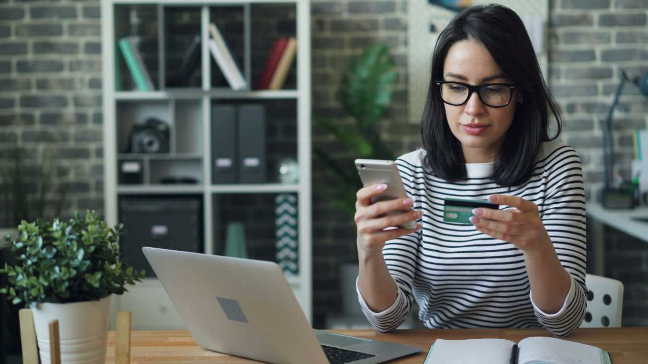 Junge Frau mit Brille macht eine online Überweisung mit der Kreditkarte und verwendet dafür Laptop und Handy