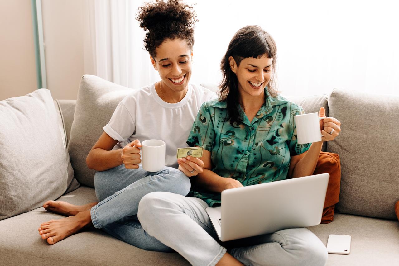 Zwei junge Frauen sitzen auf einem grauen Sofa. Beide halten eine Kaffeetasse in der Hand. Die rechte der beiden hat ihren Laptop am Schoss, die linke hält eine Kreditkarte in der Hand.