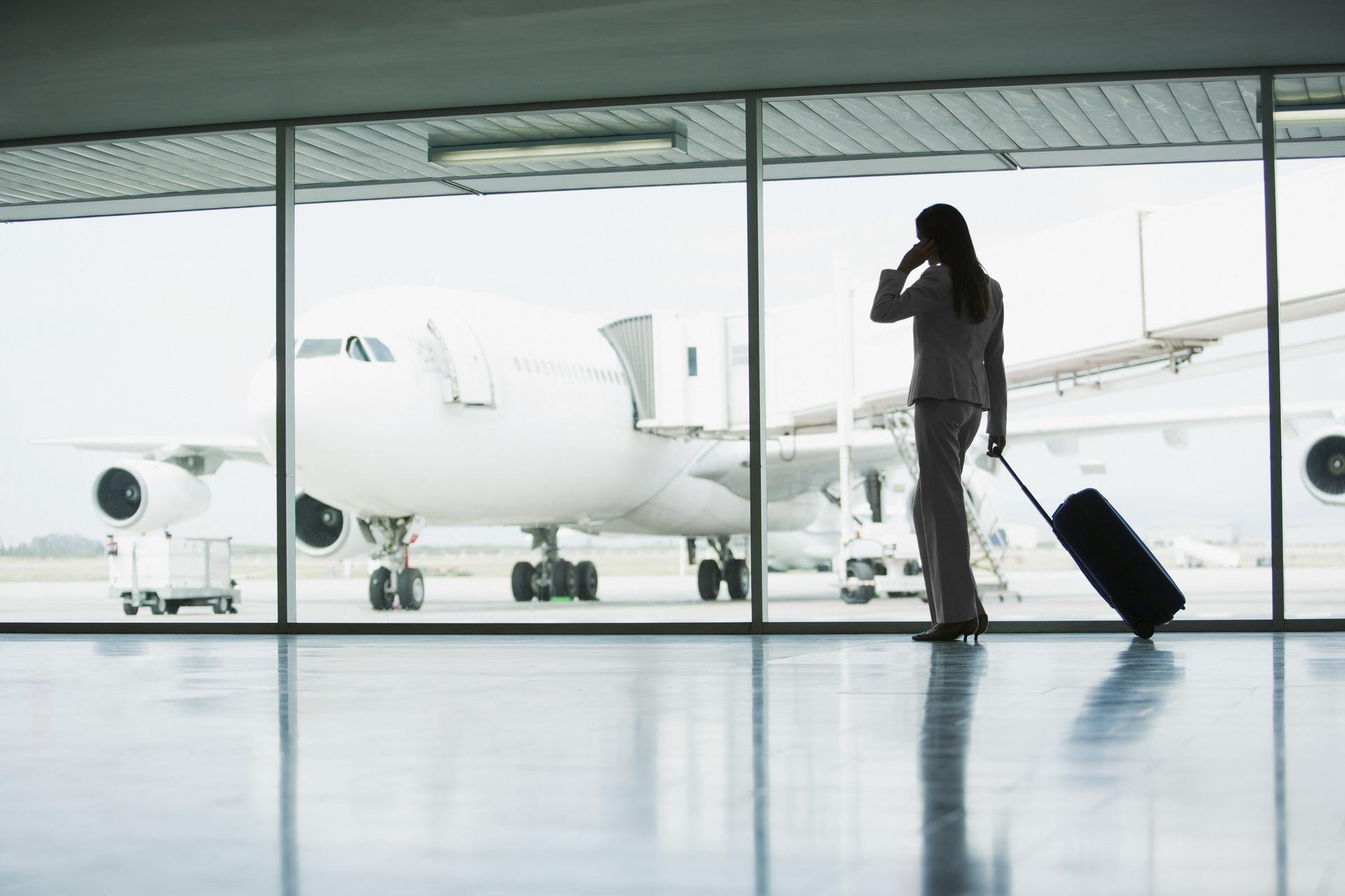 Flughafengate, eine Frau in Businessanzug mit Trolley blickt durch die verglaste Scheibe auf ein Flugzeug in Parkposition.