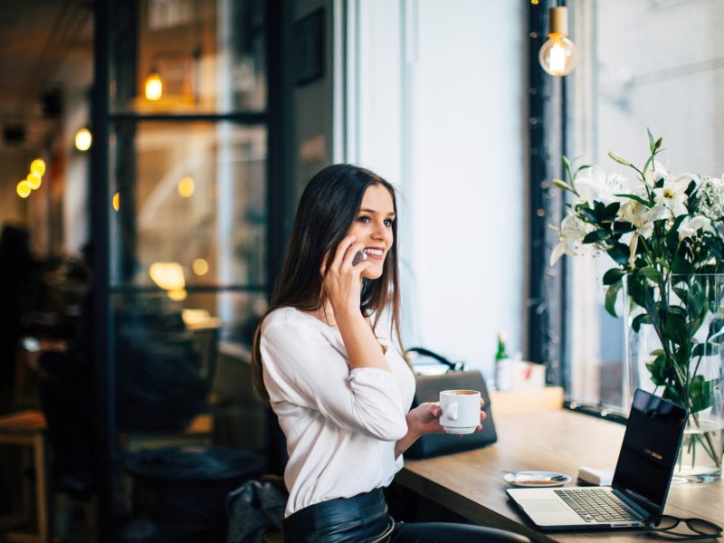 Junge dunkelhaarige Frau in weißem Pulli sitzt an einem Schreibtisch vor ihrem Laptop. Sie hält ihr Handy mit der rechten Hand ans Ohr, in der linken Hand hält sie eine Kaffeetasse. Auf dem Tisch steht ein großer Strauß weißer Lilien.