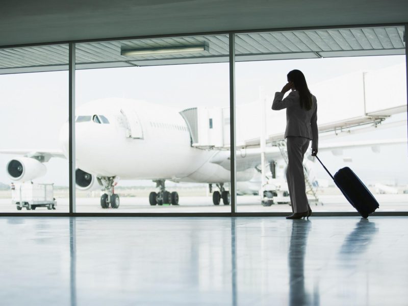Flughafengate, eine Frau in Businessanzug mit Trolley blickt durch die verglaste Scheibe auf ein Flugzeug in Parkposition.