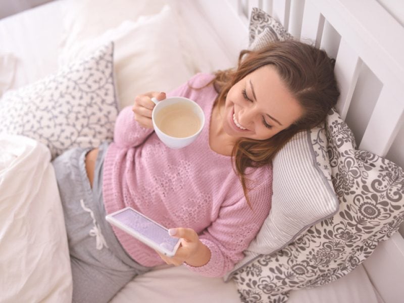 Frau liegt mit einem Kaffee in der rechten und einem Tablet in der Linken Hand im Bett