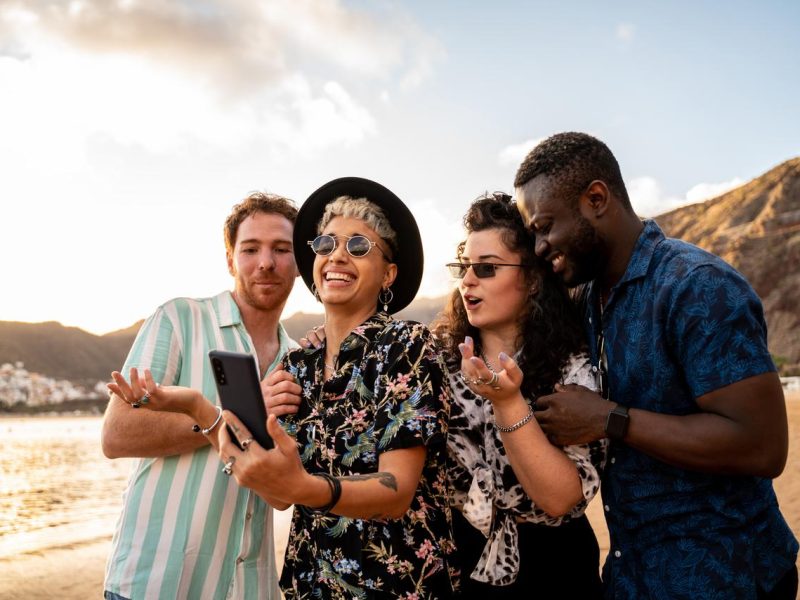 Vier Freunde machen ein Selfie am Strand. Sie lächeln und haben Spaß. Sonne, Sand und Meer im Hintergrund.