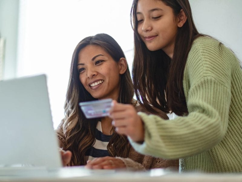 Zwei junge Frauen mit langen, dunklen Haaren schauen auf den Bildschirm eines Laptops, eine sitzend, eine stehend. Die Stehende hält eine Kreditkarte in der Hand.