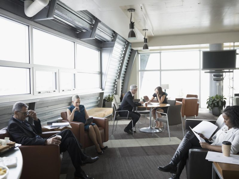 Flughafenlounge, ein Mann und zwei Frauen sitzen in bequemen Ledersesseln und sind in ihre Lektüre vertieft, an einem runden Tisch sitzen ein Mann und eine Frau und unterhalten sich.