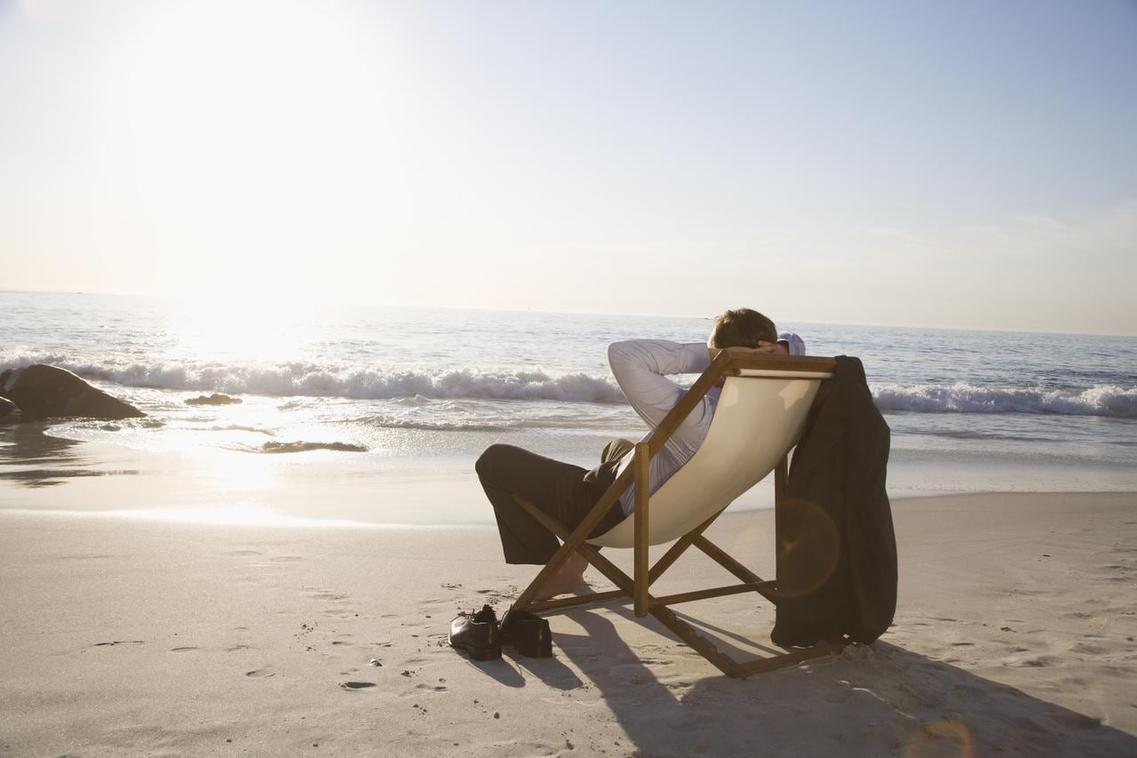 Liegestuhl am Strand, ein Mann relaxt darin, hat Schuhe und Jackett ausgezogen und blickt aufs Meer. 