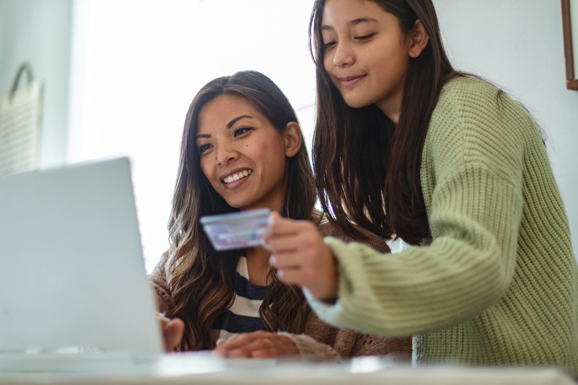 Zwei junge Frauen mit langen, dunklen Haaren schauen auf den Bildschirm eines Laptops, eine sitzend, eine stehend. Die Stehende hält eine Kreditkarte in der Hand.