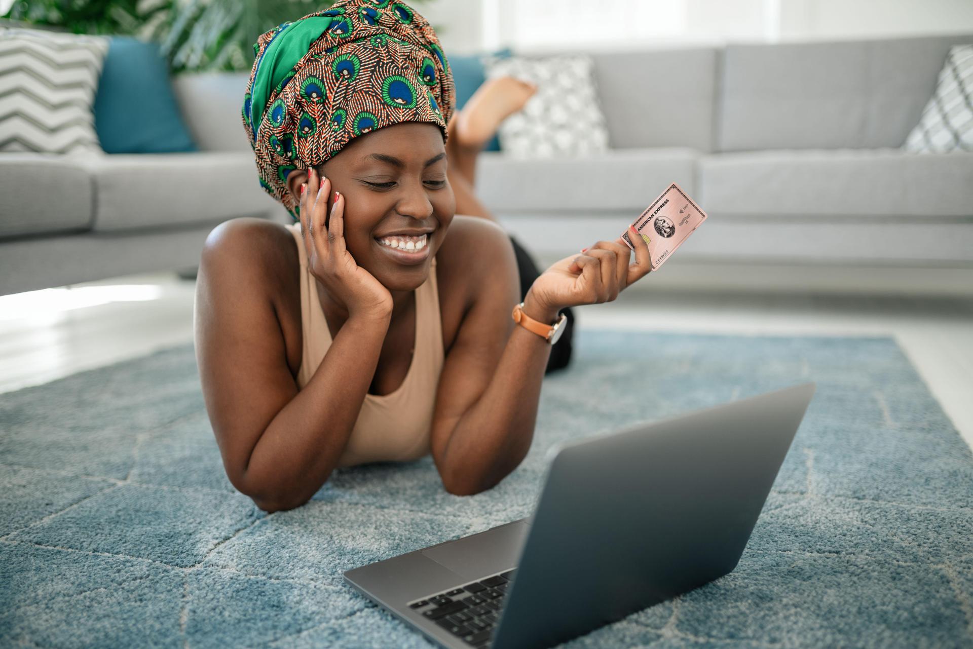 Eine junge, dunkelhäutige Frau liegt bäuchlings auf einem grauen Teppich, vor ihr ein Laptop. Sie lächelt und hält eine AMEX Karte in der linken Hand.