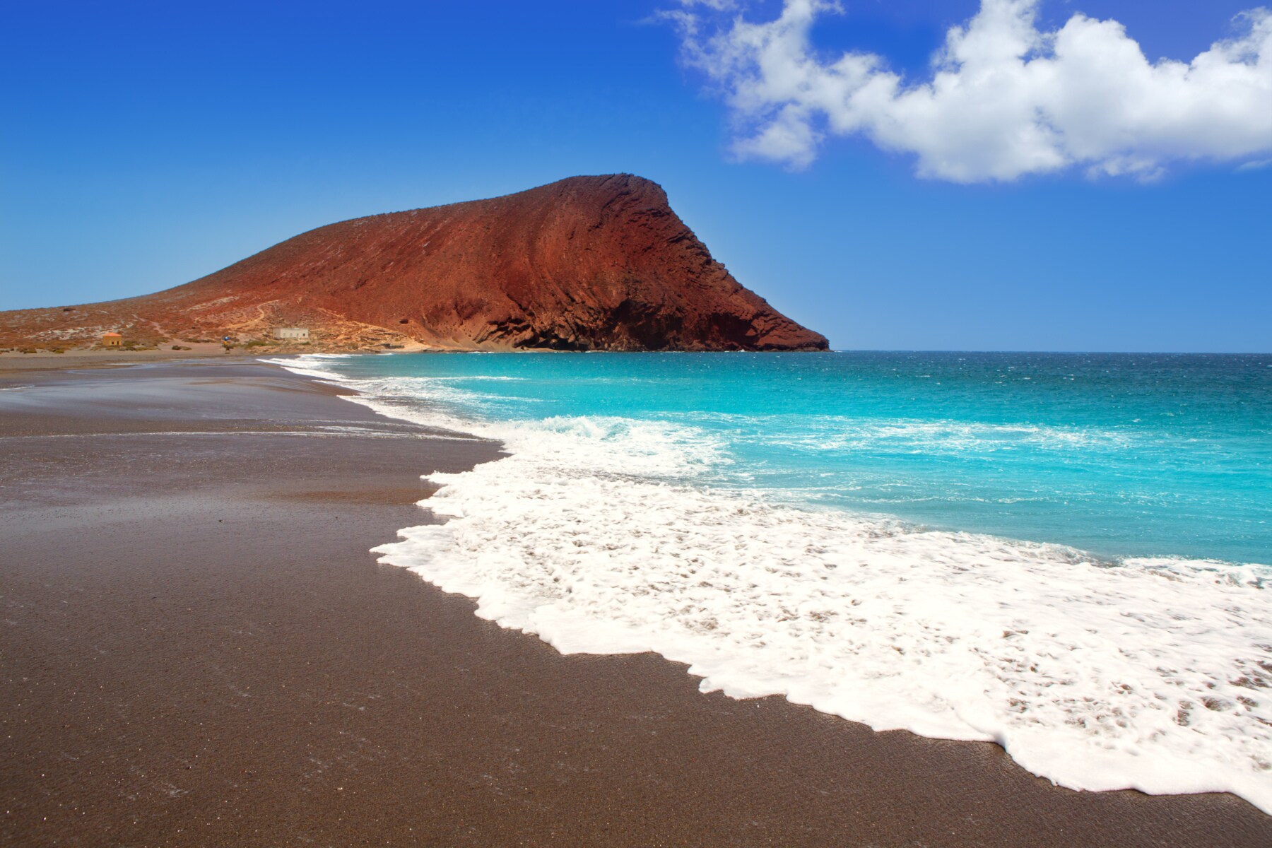 Weitläufiger Strand mit dunklem Sand und einem roten Felsen