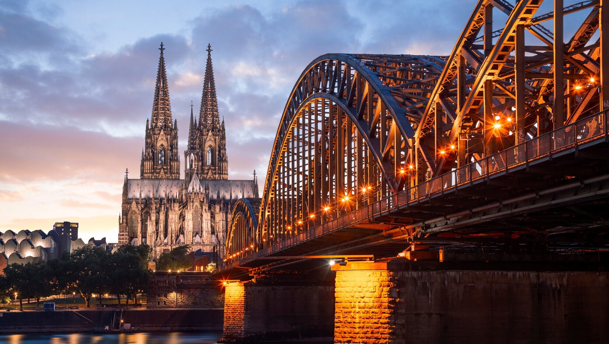 Hinter einer Brücke thront der angeleuchtete Kölner Dom in der Dämmerung