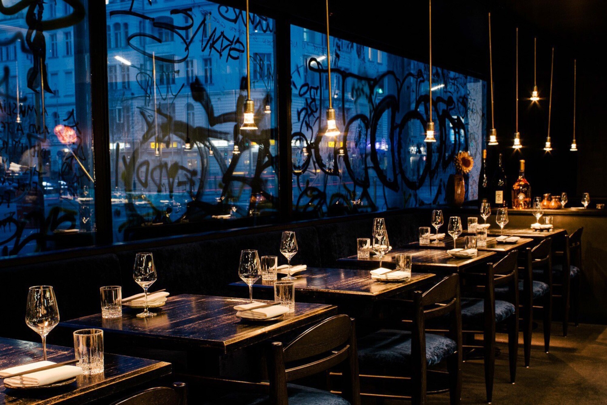 Beleuchtete, eingedeckte Holztische in einem modernen Restaurant vor einer Fensterfront mit Graffiti bei Nacht.