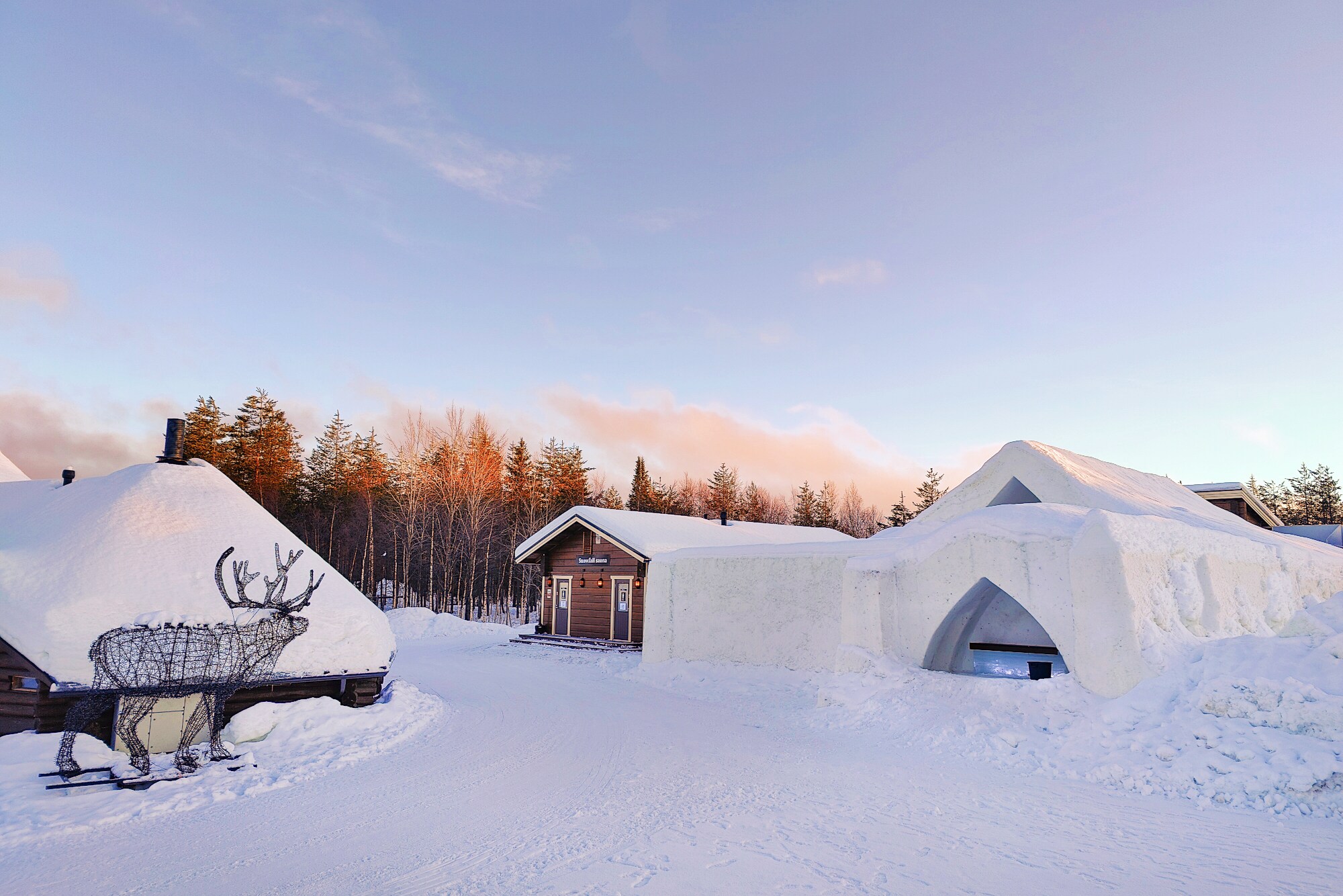 Mehrere Gebäude in einer Schneelandschaft und ein aus Ästen gebauter Hirsch.