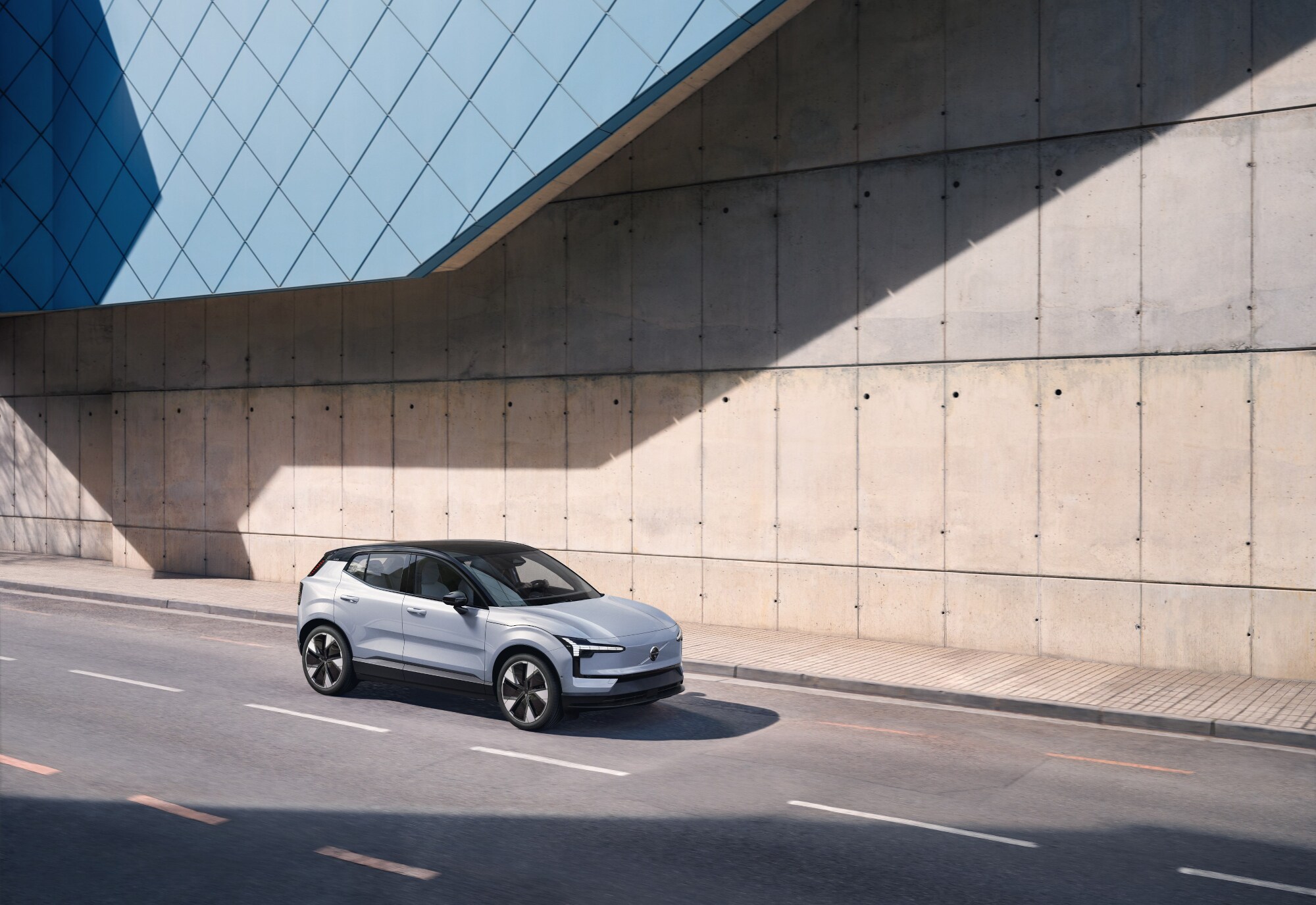 Ein silberfarbener Volvo EX30 fährt auf einer Straße an einer Betonwand vorbei.