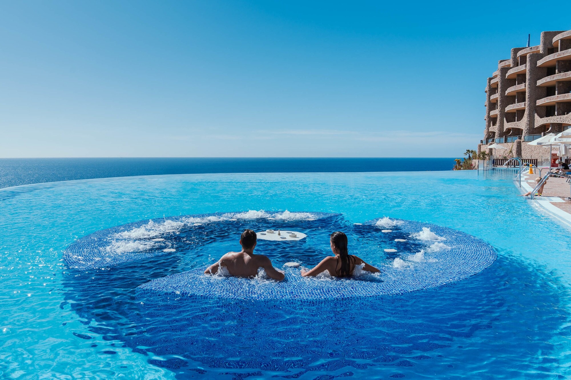 Rückansicht zweier Personen in einem großen Hotelpool mit Jacuzzi am Meer. Rückansicht zweier Personen in einem großen Hotelpool mit Jacuzzi am Meer.