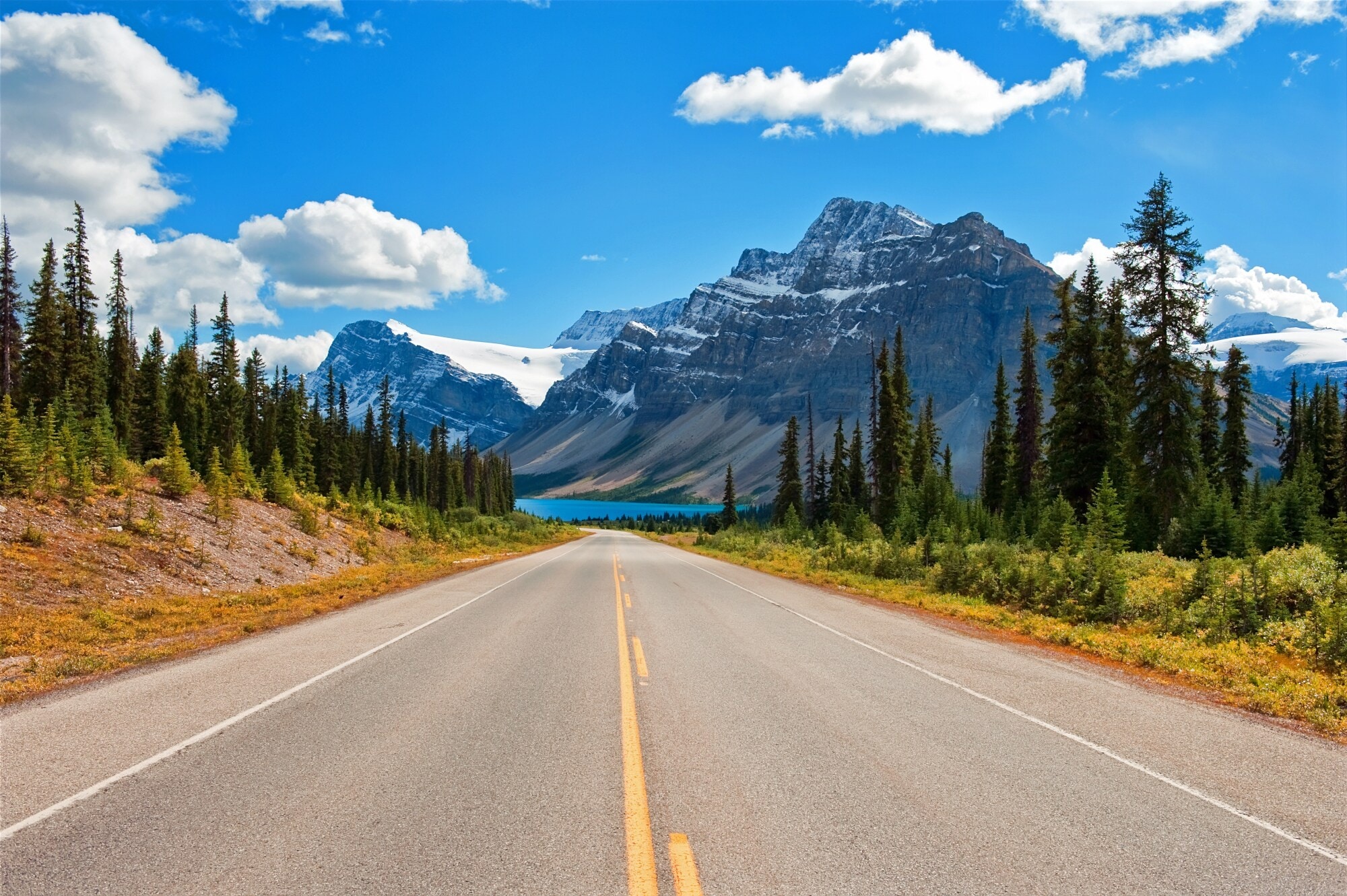 Ein Highway, umringt von Nadelbäumen, im Hintergrund schneebedeckte Berge und ein türkisblauer See. Ein Highway, umringt von Nadelbäumen, im Hintergrund schneebedeckte Berge und ein türkisblauer See.