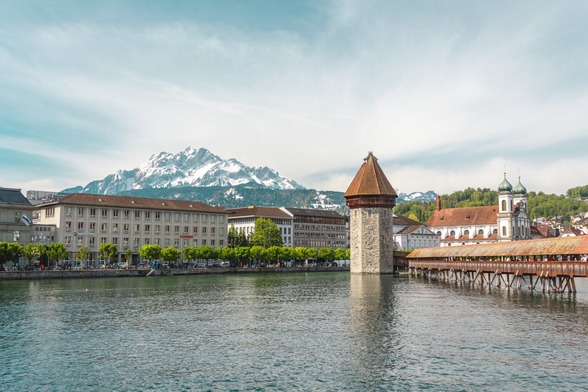 Eine Holzbrücke, ein Wasserturm, eine Stadtansicht und Berge im Hintergrund.