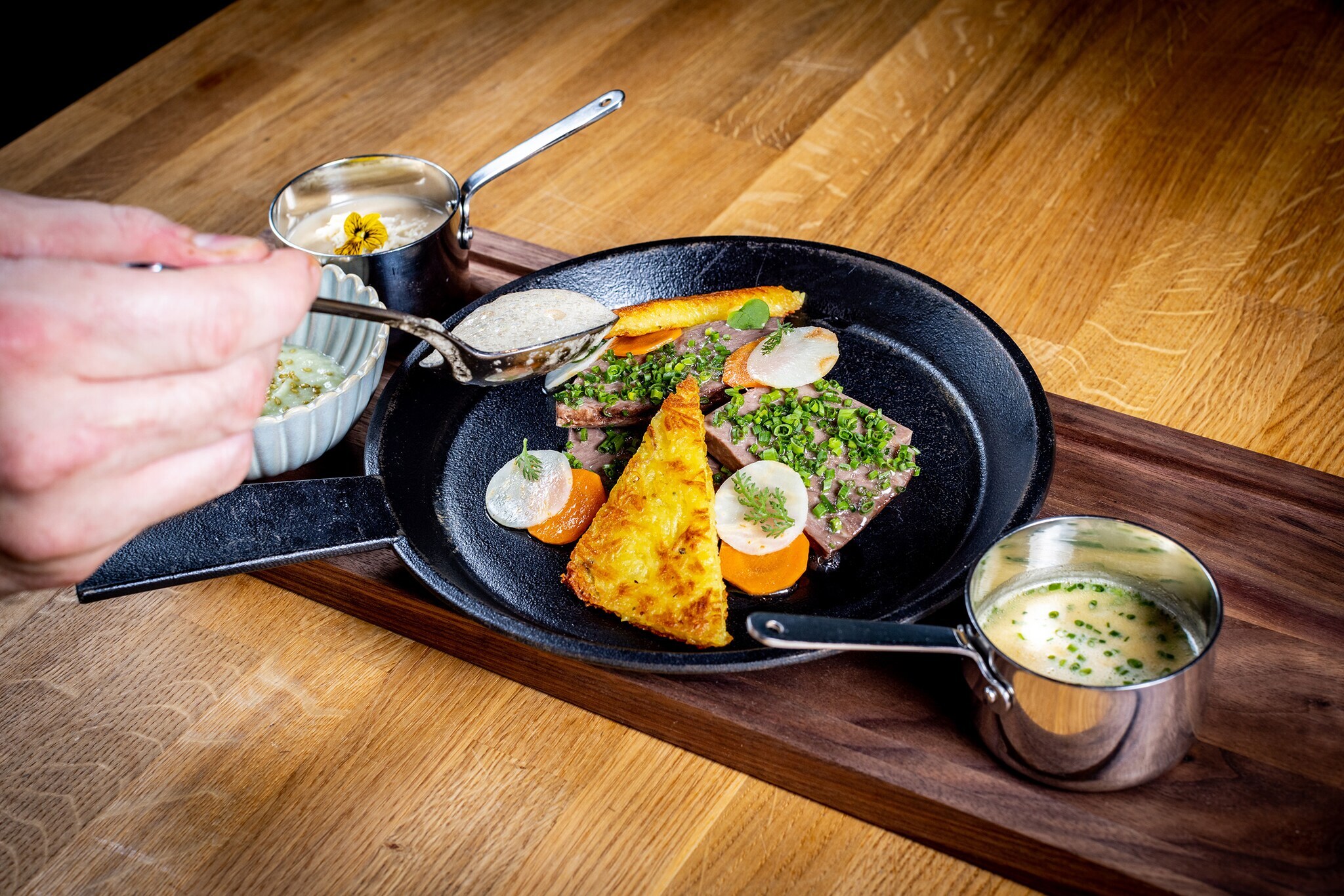 Ein Fleischgericht mit Kartoffelrösti wird in einer Gusseisenpfanne auf einem Holztisch angerichtet.