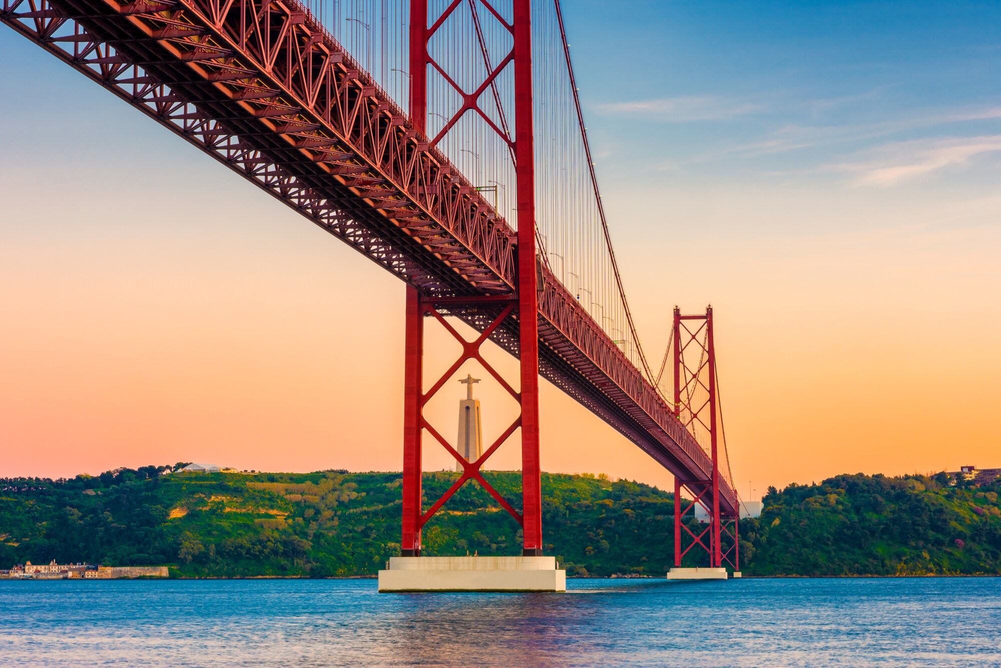Rote Hängebrücke aus Stahl über einem Fluss mit Christusstaue im Hintergrund.