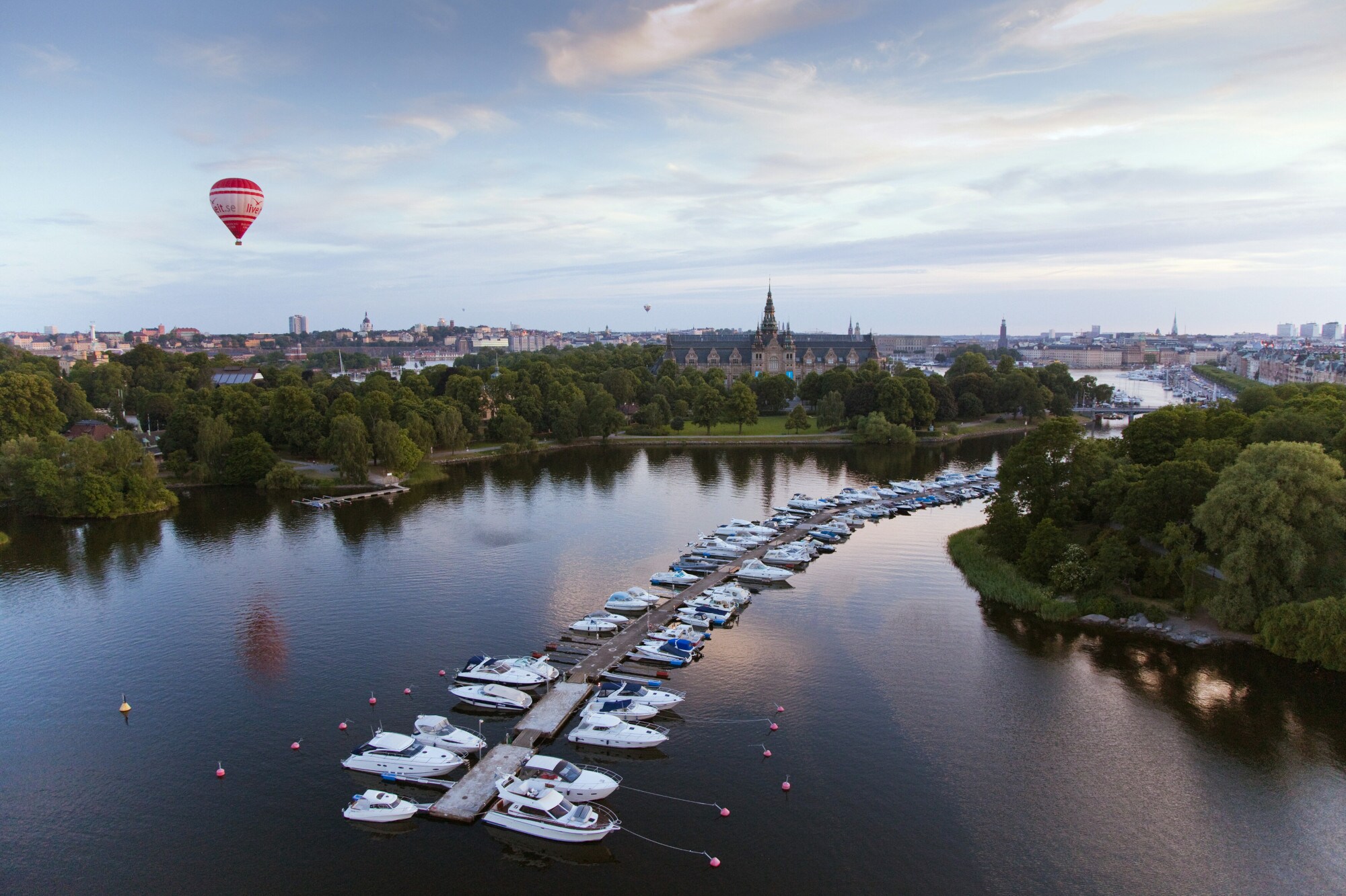 Blick über eine Stadt mit einem Fluss, Booten an einem langgezogenen Anleger und einem Heißluftballon über den Dächern. Blick über eine Stadt mit einem Fluss, Booten an einem langgezogenen Anleger und einem Heißluftballon über den Dächern.