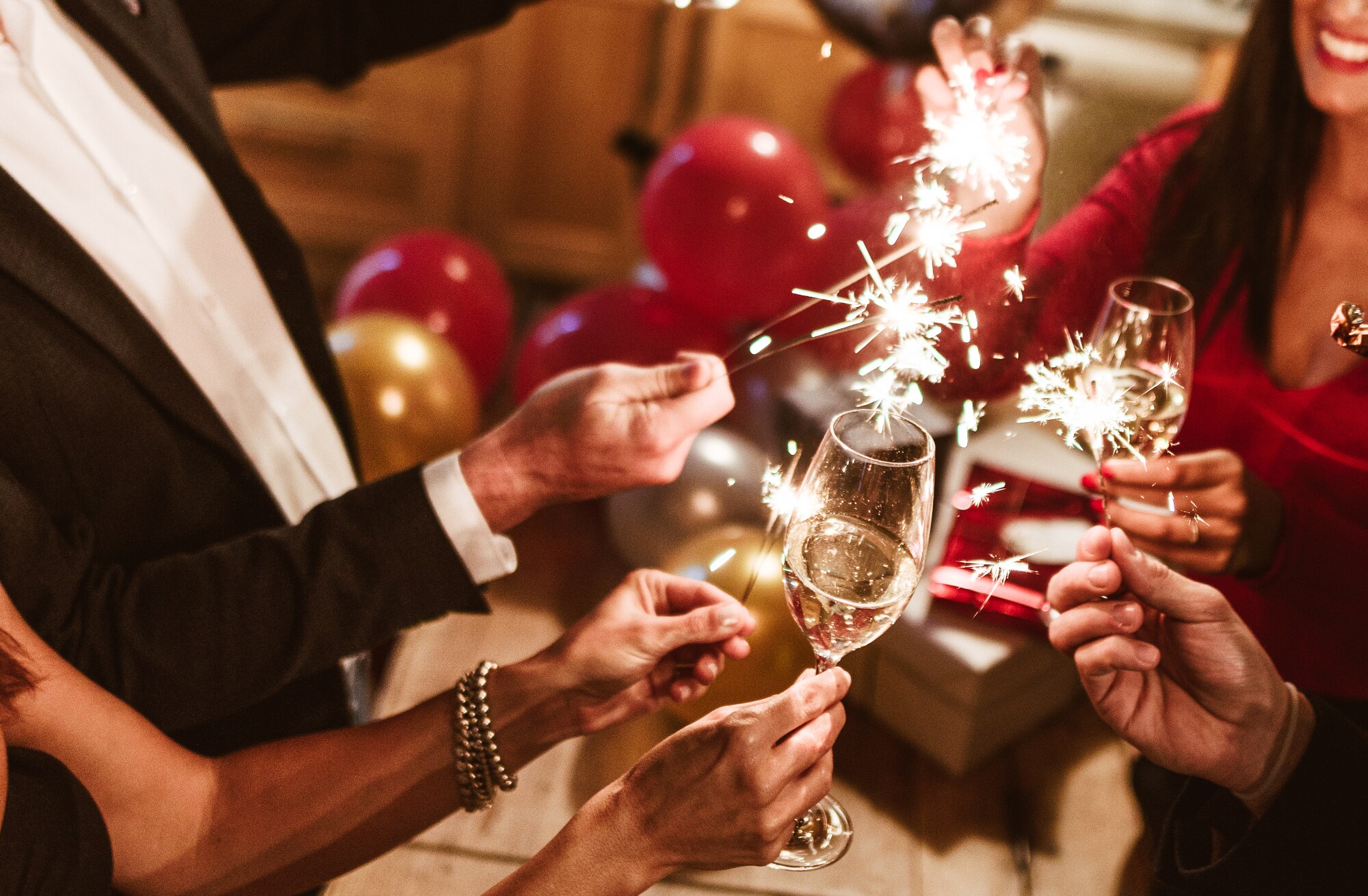 Nahaufnahme der Arme von vier edel gekleideten Personen mit Sektgläsern und Wunderkerzen im feierlichen Ambiente mit Luftballons im Hintergrund. Nahaufnahme der Arme von vier edel gekleideten Personen mit Sektgläsern und Wunderkerzen im feierlichen Ambiente mit Luftballons im Hintergrund.