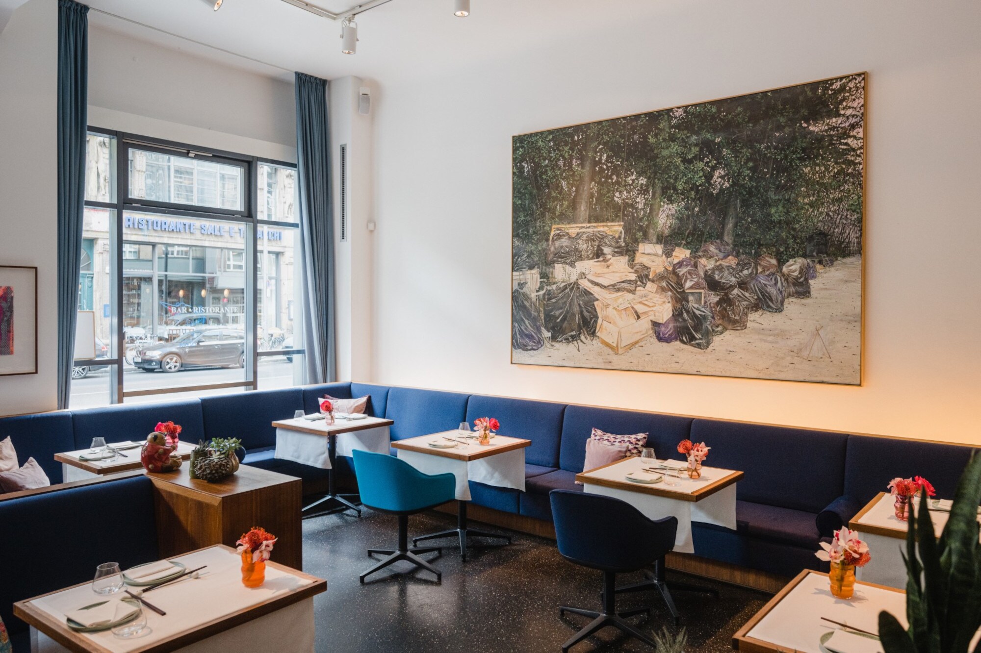 Innenraum eines hellen, modernen Restaurants mit blauen Sitzmöbeln an gedeckten Tischen und einem großen Gemälde an der Wand.