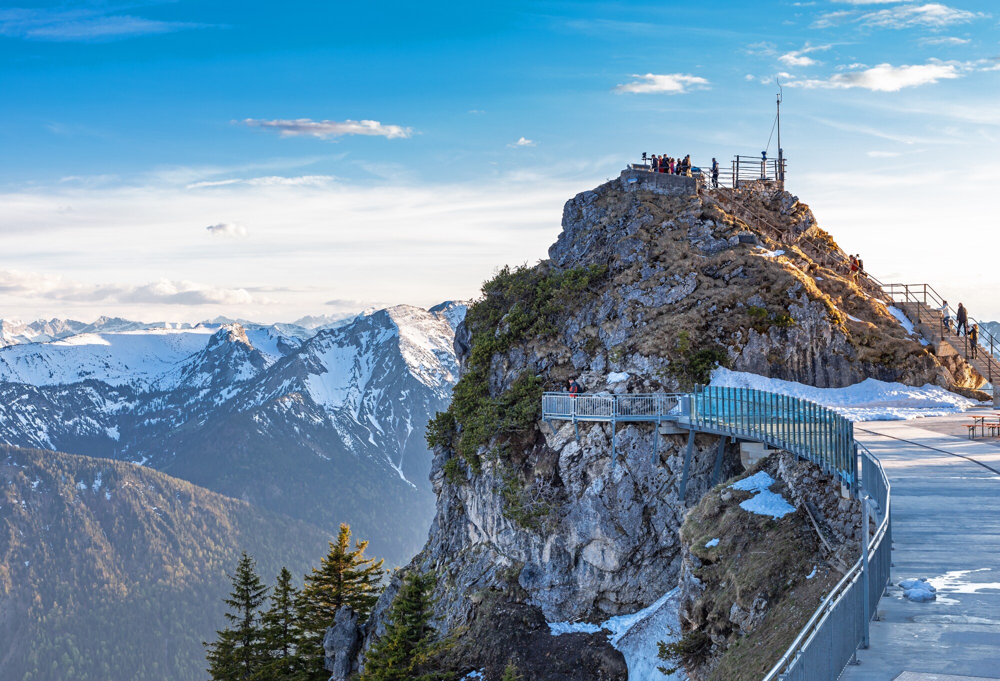 Personen auf einer Aussichtsplattform auf einem Berggipfel vor verschneitem Alpenpanorama.