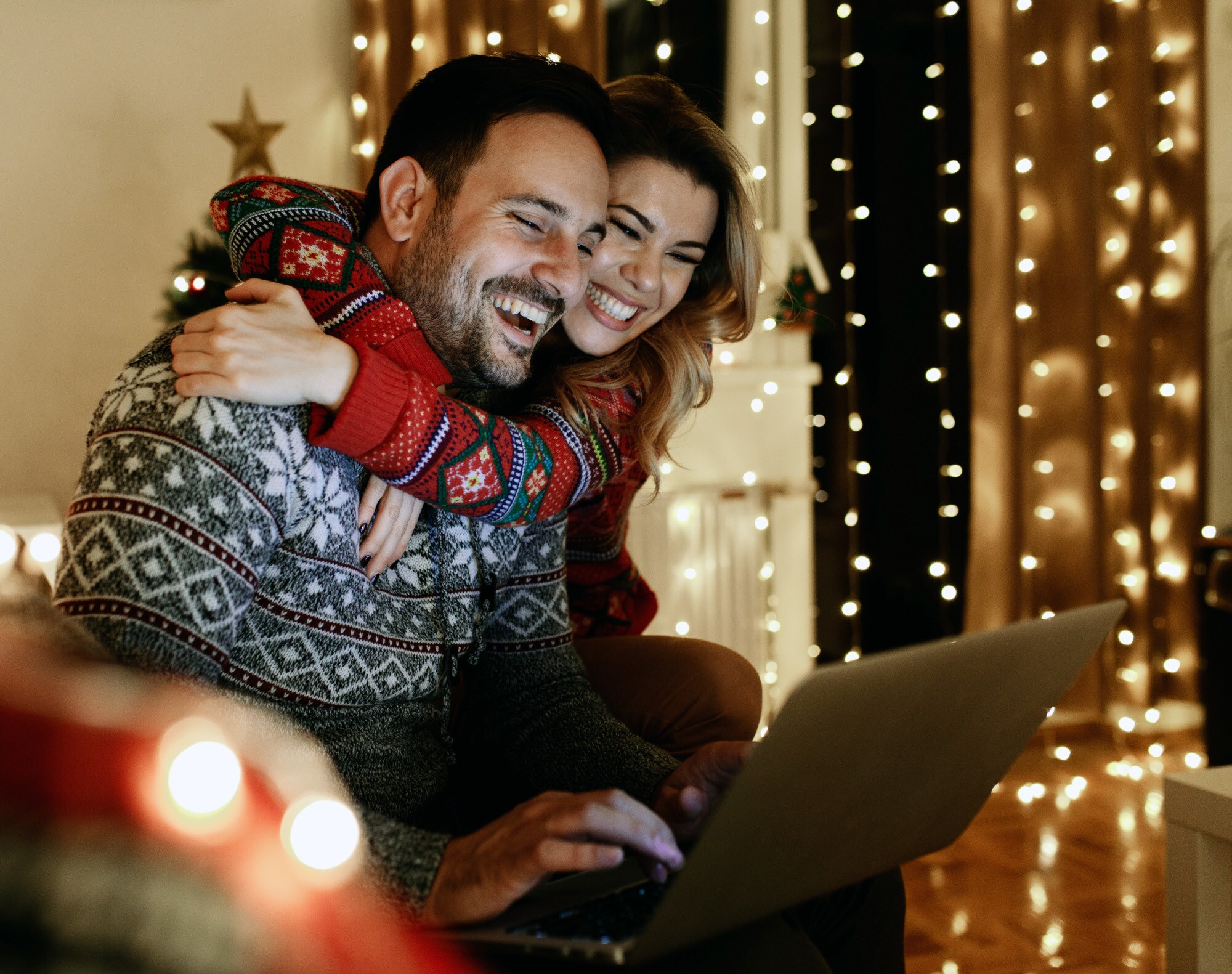 Ein lachendes Paar in Winterpullovern sitzt aneinander gekuschelt auf einem Sofa im Wohnzimmer vor einem Laptop. Ein lachendes Paar in Winterpullovern sitzt aneinander gekuschelt auf einem Sofa im Wohnzimmer vor einem Laptop.