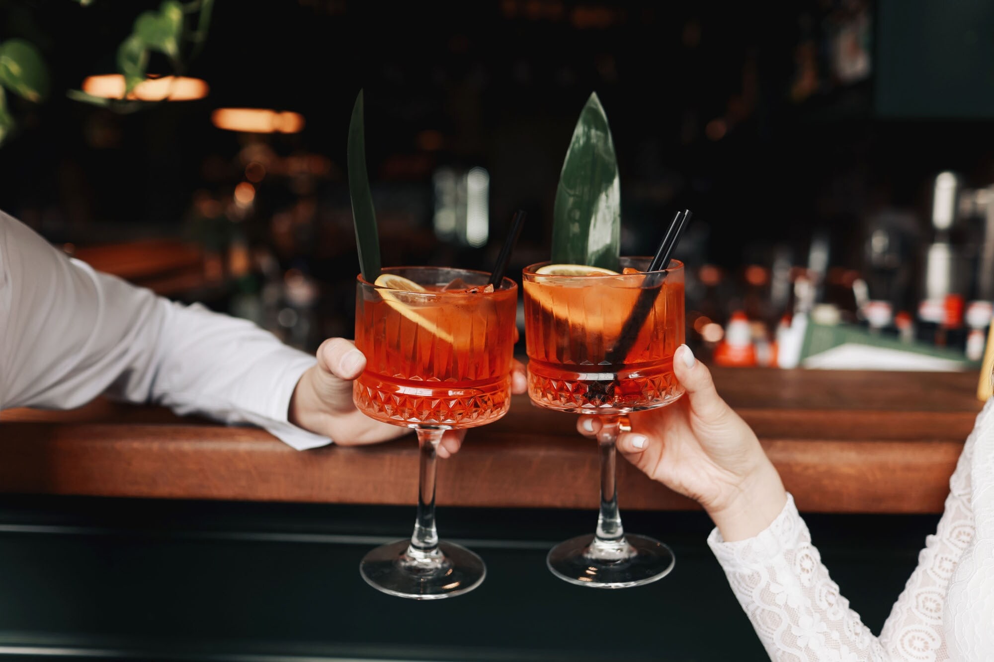 Zwei Spritz-Getränke mit Orangenscheiben in den Händen zweier Personen beim Anstoßen an einem Bartresen. Zwei Spritz-Getränke mit Orangenscheiben in den Händen zweier Personen beim Anstoßen an einem Bartresen.