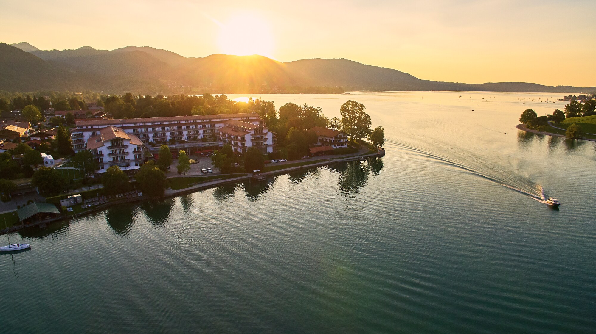 Luxuriöser Hotelkomplex an einem Seeufer, umgeben von Bäumen vor einer bewaldeten Hügellandschaft. Luxuriöser Hotelkomplex an einem Seeufer, umgeben von Bäumen vor einer bewaldeten Hügellandschaft.