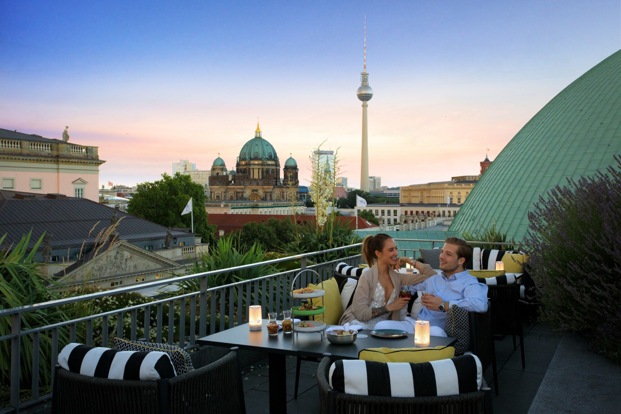 Ein junges Paar sitzt gut gelaunt an einem Tisch auf einer Dachterrasse, im Hintergrund Berliner Dom und Fernsehturm.