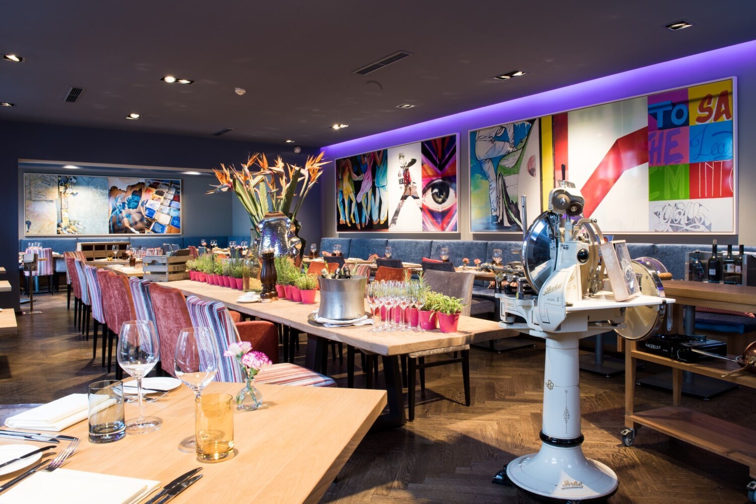 Restaurant mit einem langen, mit Pflanzen dekorierten Holztisch und einer Aufschnittmaschine mit Standfuß, umgeben von farbenfrohen Bildern an der Wand.