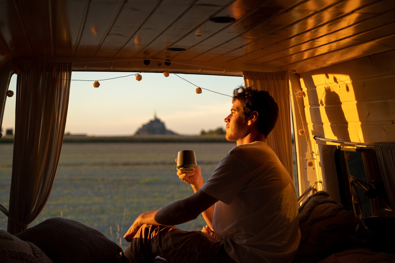 Ein junger Mann sitzt mit einem Kaffeebecher in der Hand im geöffneten Campervan im goldenen Sonnenlicht und blickt hinaus in die Natur.