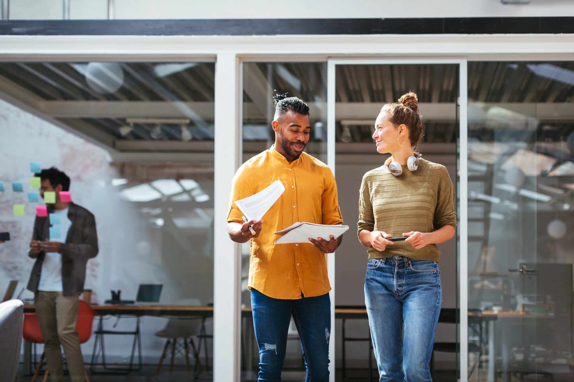 Zwei lässig gekleidete Personen stehen mit Arbeitsmaterialen vor einem offenen Büro mit einem langen Tisch. Zwei lässig gekleidete Personen stehen mit Arbeitsmaterialen vor einem offenen Büro mit einem langen Tisch.