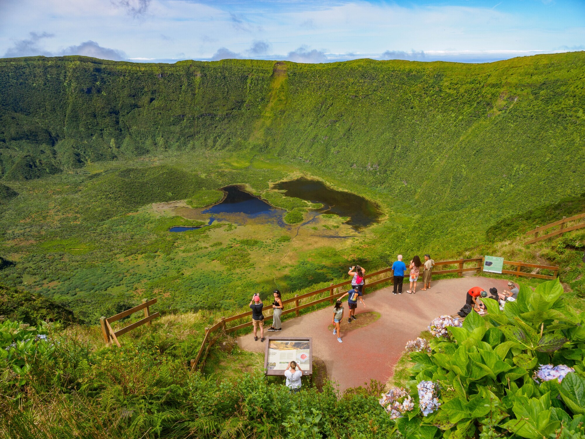 Personen auf einer Aussichtsplattform am Rande eines von Grün bedecktem Vulkankrater.
