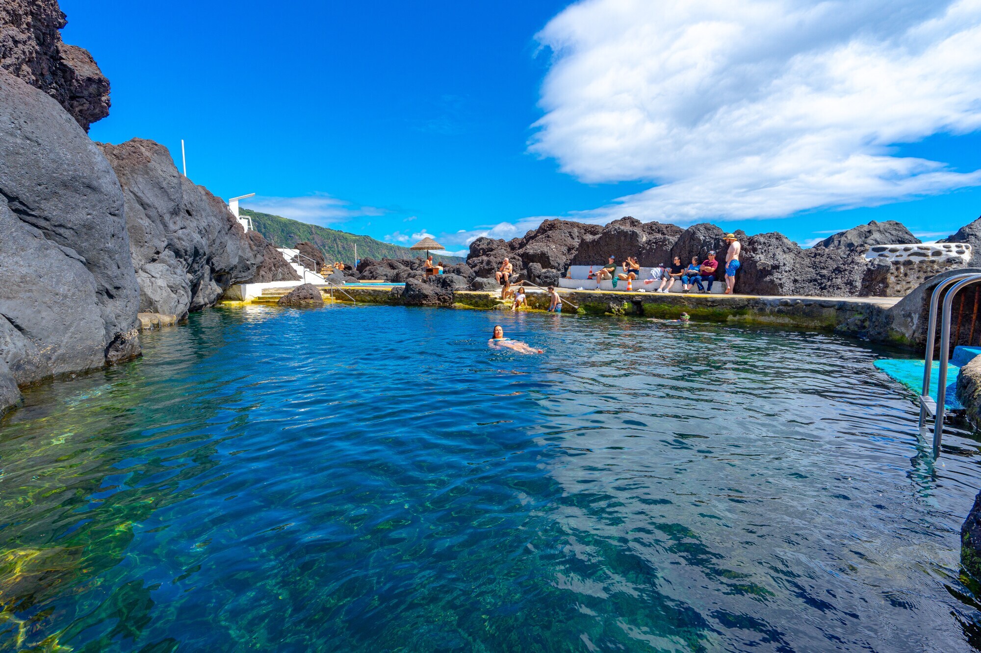 Personen an und in einem Naturschwimmbecken, umgeben von Felsbrocken.