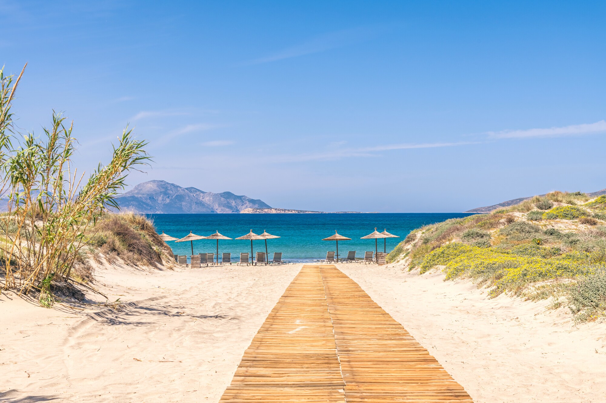 Weg durch Dünen zu einem hellen Sandstrand mit Sonnenliegen am türkisblauen Meer.