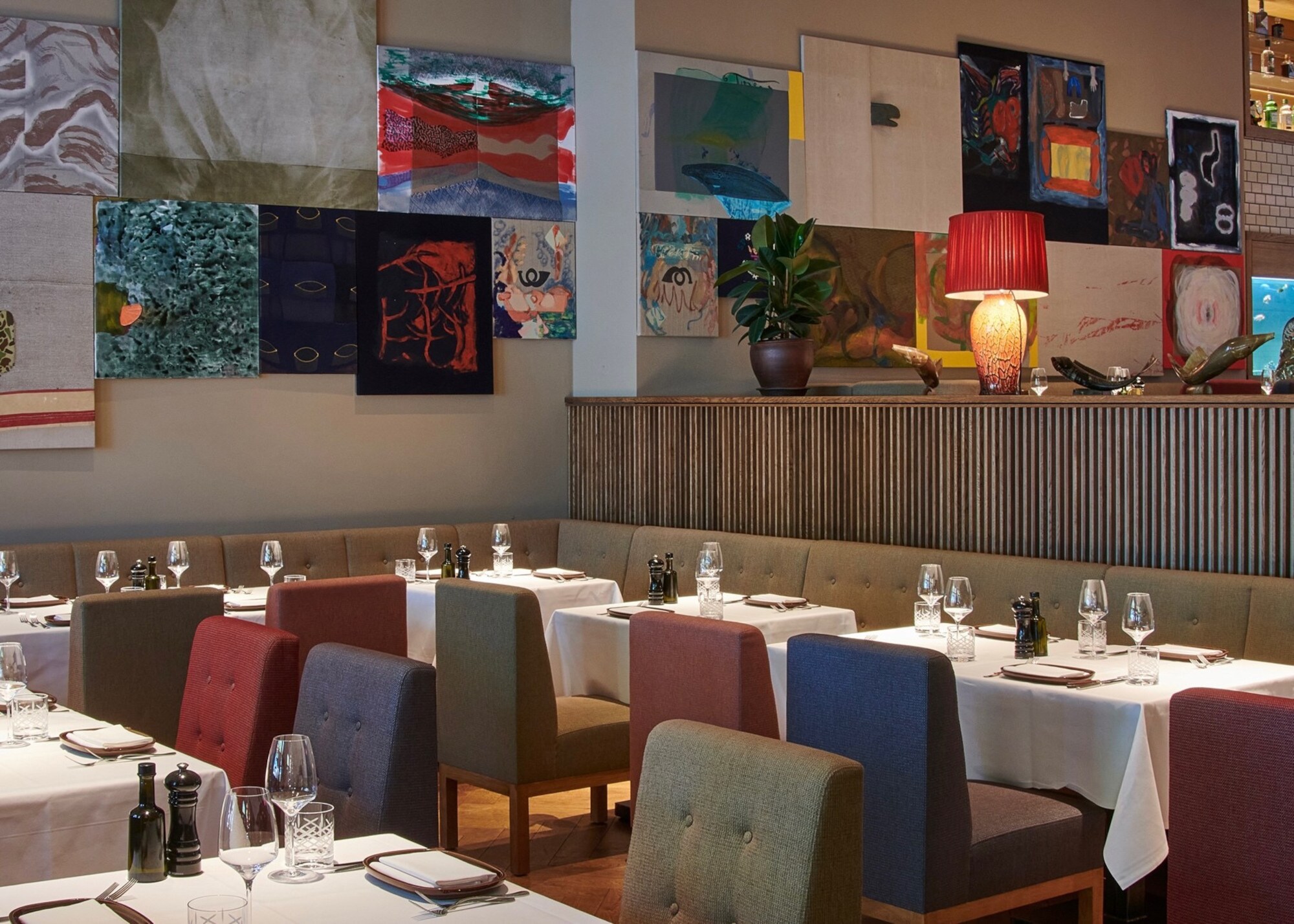 Restaurant mit Polsterstühlen an elegant gedeckten Tischen vor einer Wand mit farbenfrohen Gemälden.