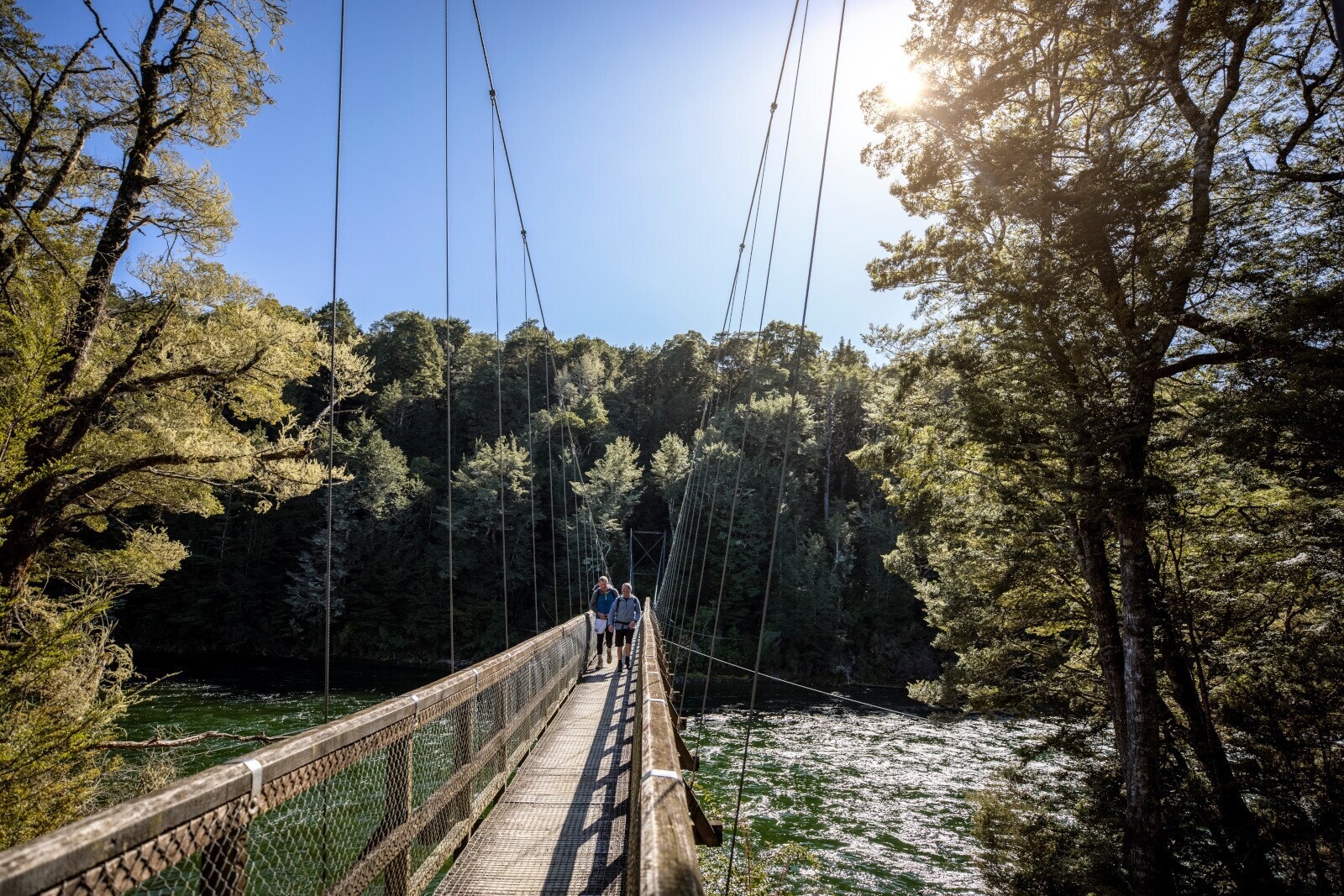 Zwei Personen laufen auf einer Hängebrücke über einen Fluss in einem Waldgebiet. Zwei Personen laufen auf einer Hängebrücke über einen Fluss in einem Waldgebiet.