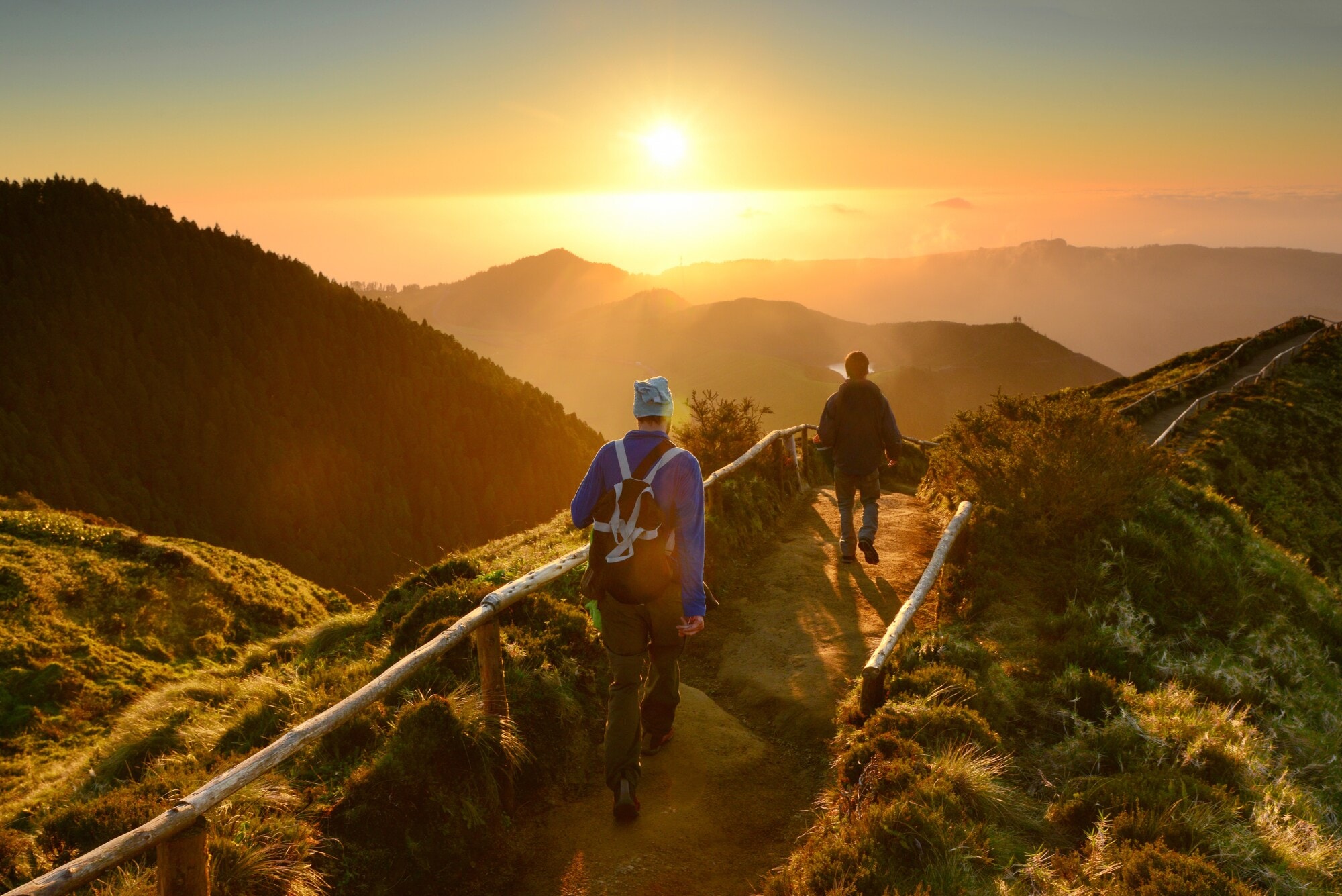 Rückansicht zweier gehender Personen auf einem Weg entlang eines Bergkamms inmitten einer grünen Gebirgslandschaft bei Sonnenuntergang. Rückansicht zweier gehender Personen auf einem Weg entlang eines Bergkamms inmitten einer grünen Gebirgslandschaft bei Sonnenuntergang.