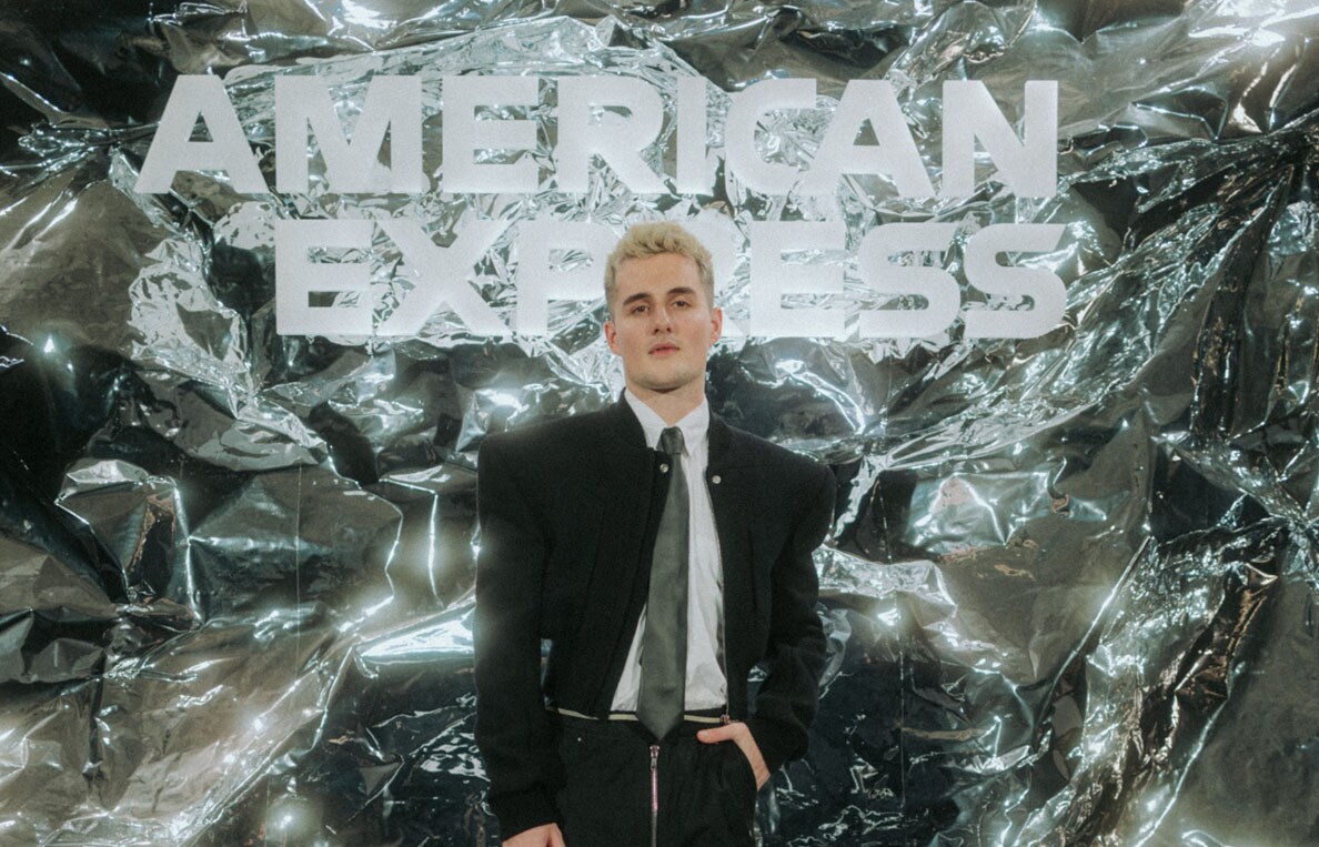 Luis Freitag von den Elevator Boys im schwarzem Anzug mit weißem Hemd und Krawatte vor einer silbernen Wand mit weißem American Express Schriftzug.