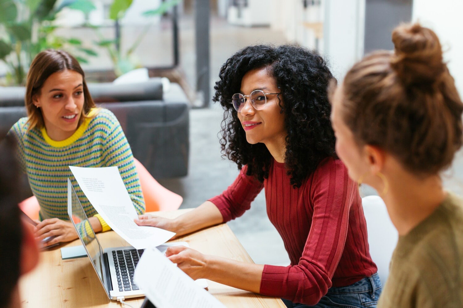 Drei junge Frauen sitzen mit Arbeitsunterlagen zusammen an einem Holztisch in einem Co-Working-Space. Drei junge Frauen sitzen mit Arbeitsunterlagen zusammen an einem Holztisch in einem Co-Working-Space.